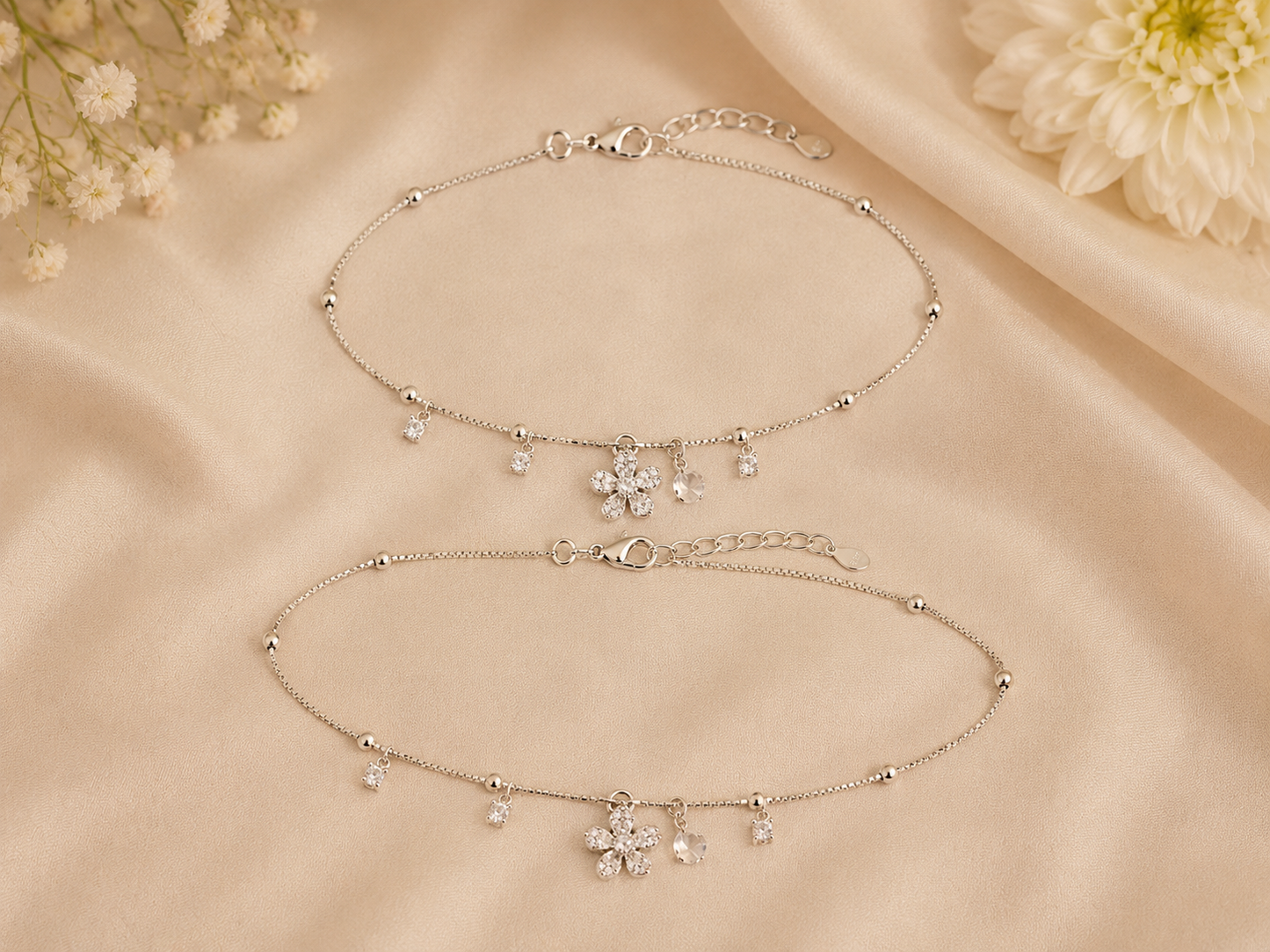 Two silver anklets with floral charms on a beige fabric background.