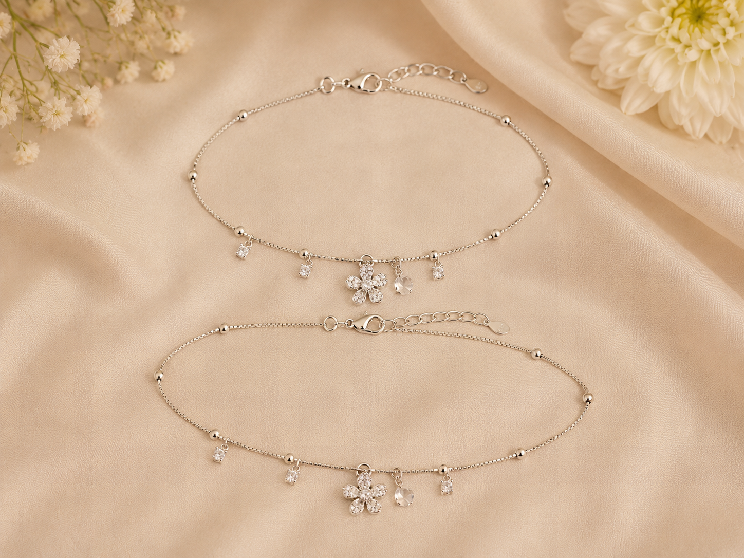 Two silver anklets with floral charms on a beige fabric background.