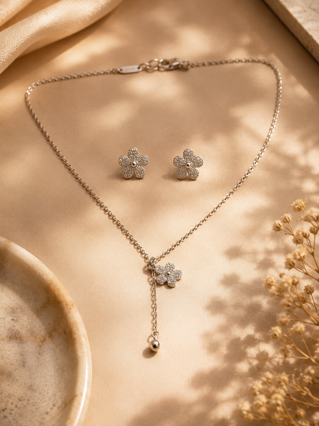 Silver necklace and earrings on a beige surface with dried flowers.